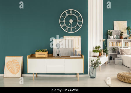 White cabinet, paintings and modern clock on a green wall with a bedroom interior in the background Stock Photo