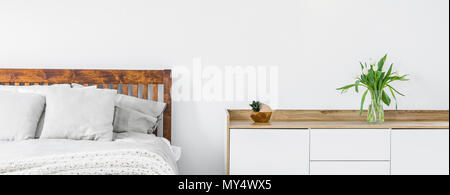 Close-up of a part of wooden bed with linen and pillows and a side cabinet with fresh cut flowers in a vase on top standing against a white wall in a  Stock Photo