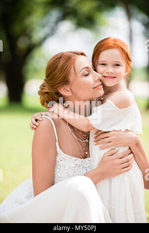 beautiful happy red haired mother and daughter in white dresses hugging at park Stock Photo