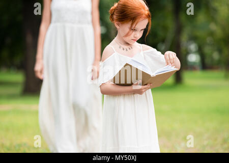 adorable red haired girl in white dress reading book while standing at park Stock Photo