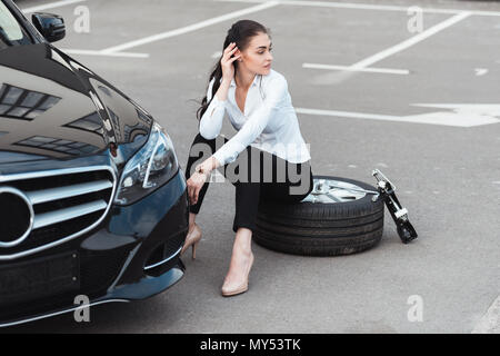Young attractive woman sitting on car tire in parking lot Stock Photo