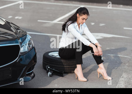 Young attractive woman sitting on car tire with lug wrench in her hand in parking lot Stock Photo