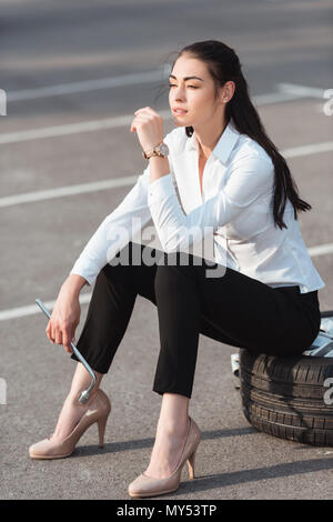 Young attractive woman sitting on car tire with lug wrench in her hand in parking lot Stock Photo