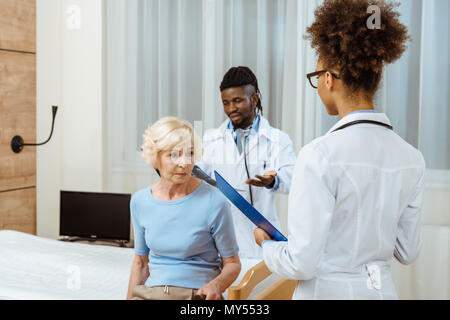 Elderly woman sitting on a hospital bed with two young doctors with clipboard and x-ray photo standing over her Stock Photo