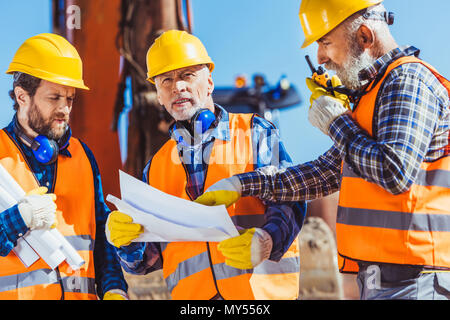 Three workers examining building plans and talking on portable radio at construction site Stock Photo