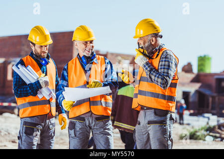 Three workers in hardhats examining building plans and talking on portable radio at construction site Stock Photo
