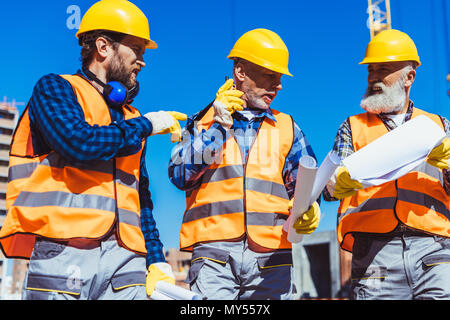 Three workers in reflective vests and hardhats walking together and discussing building plans Stock Photo