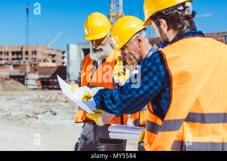 Three workers in reflective vests and hardhats standing at construction site and examining building plans Stock Photo
