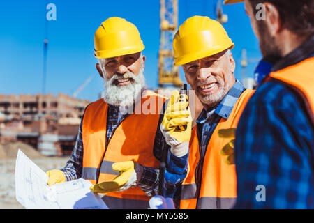 Two workers in reflective vests and hardhats examining building plans and talking on portable radio at construction site Stock Photo