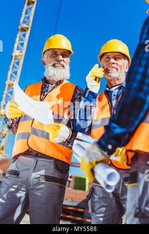 Two workers in hardhats and protective vests standing at construction site with building plans and portable radio Stock Photo