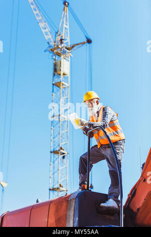 Smiling worker in reflective vest and hardhat using digital tablet while standing on top of excavator cabin Stock Photo