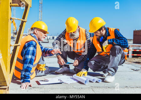 Three construction workers sitting on concrete at construction site, discussing building plans Stock Photo