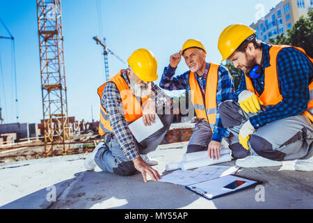 Three construction workers in uniform sitting on concrete at construction site, examining building plans Stock Photo