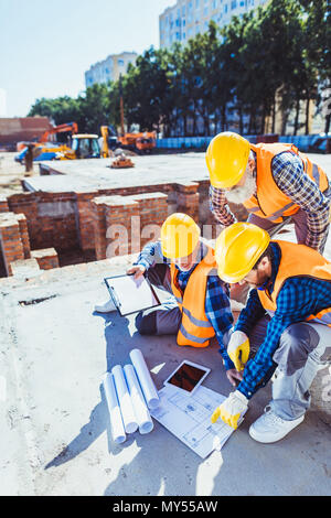 Three construction workers in hardhats sitting on concrete at construction site, examining building plans Stock Photo
