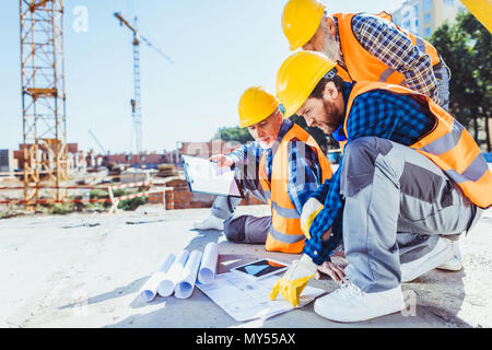 construction workers in uniform sitting on concrete at construction site, discussing building plans Stock Photo