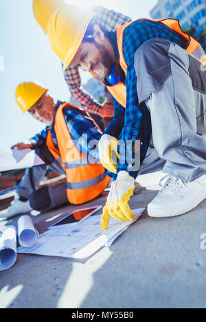 Smiling construction workers in hardhats sitting on concrete at construction site, examining building plans Stock Photo
