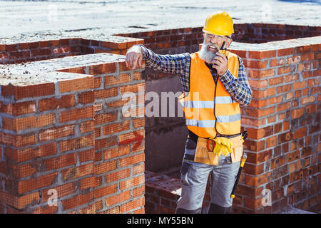 Construction worker in reflective vest and hardhat leaning on unfinished wall and talking on portable radio Stock Photo