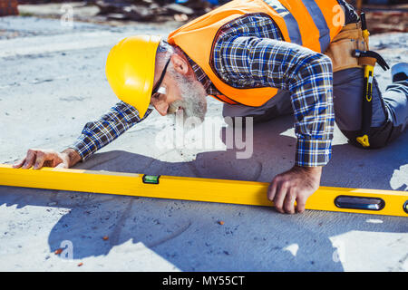Bearded worker in reflective vest and hardhat working with spirit level at construction site, leveling the floor Stock Photo