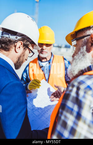 Workers in protective uniform discussing building plans with businessman at construction site Stock Photo