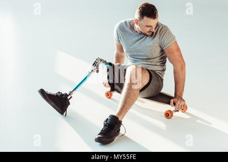 young man with leg prosthesis sitting on skateboard isolated on white Stock Photo