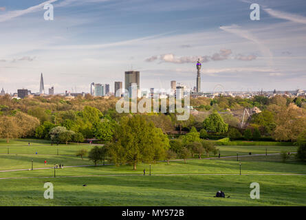 London, England, UK - April 26, 2018: People sit and walk on the slopes of Primrose Hill in Regent's Park, with the skyline of central London behind. Stock Photo