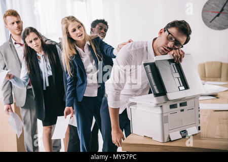 exhausted businessman sleeping on copier while his colleagues standing in queue behind him Stock Photo