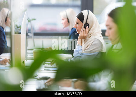 selective focus of smiling arabic call center operator in hijab in headset working on computer at workplace in office Stock Photo
