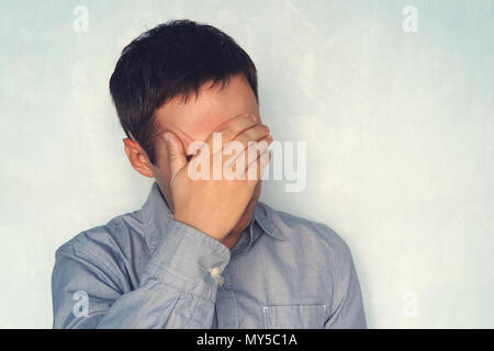 a young guy in a blue shirt is holding his head. the concept of fatigue. Close your eyes to the problem. Portrait of a handsome man covering his eyes  Stock Photo