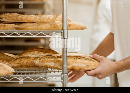 cropped shot of handsome baker putting fresh bread on shelf at baking manufacture Stock Photo