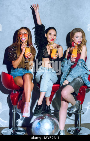 beautiful fashionable multiethnic women sitting on bar stools and drinking cocktails at party Stock Photo