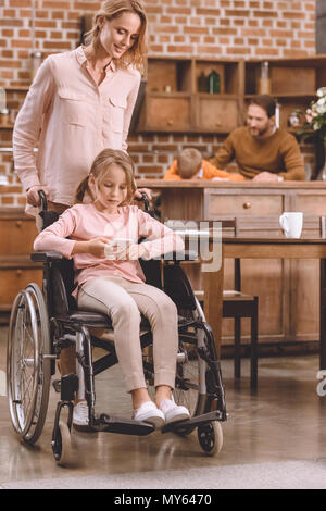 smiling mother looking at disabled daughter sitting in wheelchair and using smartphone at home Stock Photo