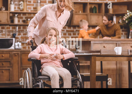 mother looking at smiling disabled daughter sitting in wheelchair and using smartphone at home Stock Photo