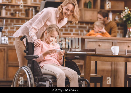 happy mother looking at smiling disabled daughter sitting in wheelchair and using smartphone at home Stock Photo