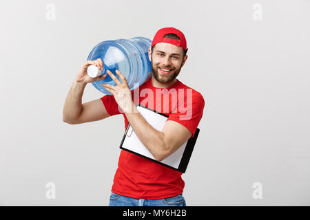 Handsome young delivery worker in red uniform is holding a bottle of water, looking at camera and smiling, on gray background Stock Photo