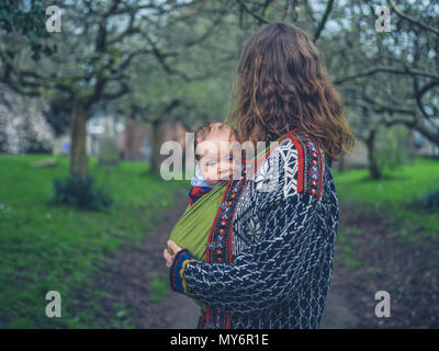 A young mother is standing in the park with her baby in a carrier sling Stock Photo