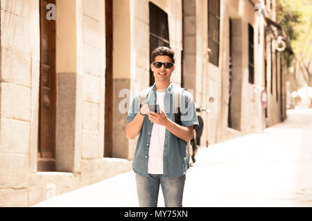 young happy student man in jeans denim clothes wearing sunglasses texting on his smart mobile phone and smiling, outside on the street of a modern eur Stock Photo