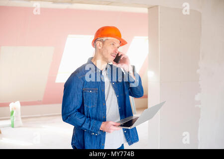 Construction man making call and using his laptop while standing at construction site. Stock Photo