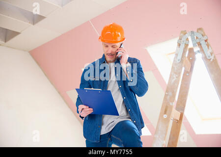 Portrait of construction worker using his mobile phone and consulting with somebody while standing at the ladder. Stock Photo