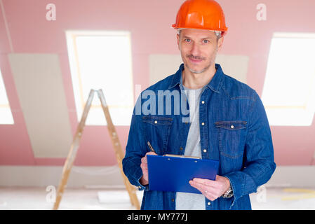 Middle aged handyman wearing hardhat and holding clipboard in his hand while standing at construction site. Stock Photo