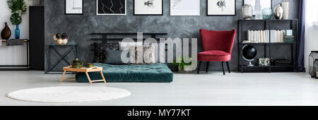 Red armchair standing next to metal rack with books and decorations in dark grey bedroom interior with green mattress and white rug Stock Photo