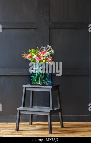 Bouquet of colorful flowers in blue vase on black stool in dark minimal interior with wooden wall Stock Photo