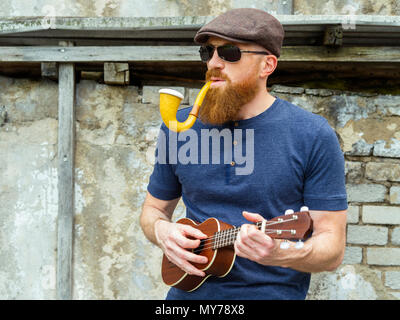 Photo of a bearded man in his thirties smoking a large pipe and playing a ukulele. Stock Photo