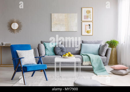 Real photo of a blue armchair standing next to a white table in a modern living room interior with a grey sofa and posters Stock Photo