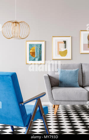 Real photo of a blue armchair standing in front of a grey couch in living room interior with posters in gold frames and checkered floor Stock Photo