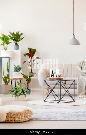 Real photo of a beige living room interior with a sofa standing behind a black table, under a lamp and next to plants Stock Photo