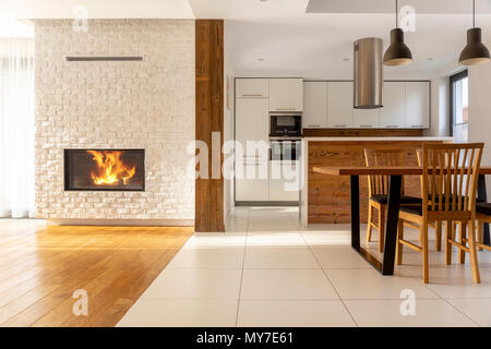 Fireplace in spacious white house interior with wooden chairs at table near kitchen. Real photo Stock Photo
