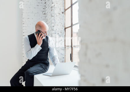 Businessman sitting on windowsill using laptop and smartphone Stock Photo