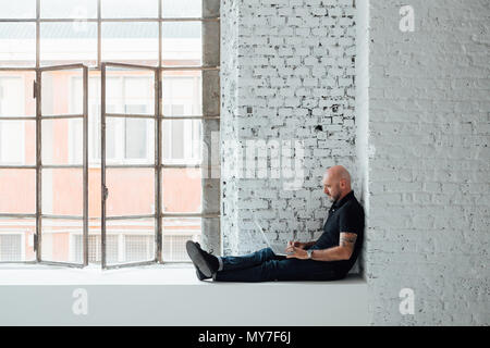 Man sitting on windowsill using laptop, full length, side view Stock Photo
