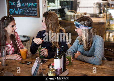 Three young female friends with vegetable juice chatting in cafe Stock Photo