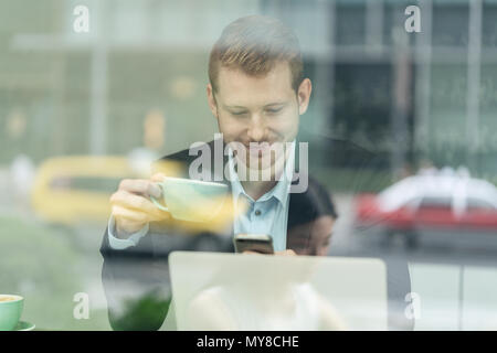 Businessman sitting in cafe, drinking coffee, using smartphone, viewed through window Stock Photo
